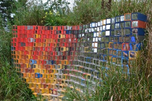 A colorful, abstract wall made of small, reflective squares, surrounded by tall grass and greenery.