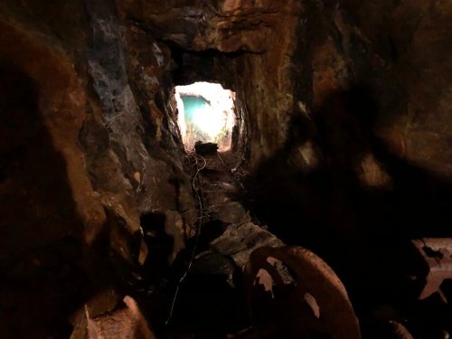 A dimly lit mine tunnel with rocky walls, leading to a bright opening in the distance. Shadows cast on the ground.
