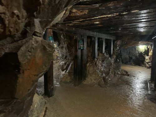 Dimly lit underground mine tunnel with wooden supports and lanterns illuminating the rocky walls.