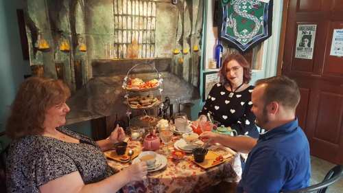 Three people enjoy a tea party at a table adorned with pastries and drinks, set against a cozy, artistic backdrop.