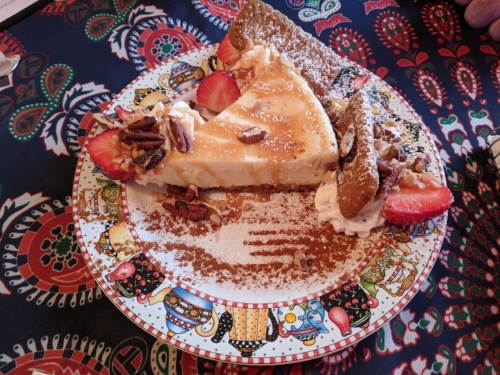 A slice of cheesecake topped with strawberries, nuts, and whipped cream on a decorative plate.