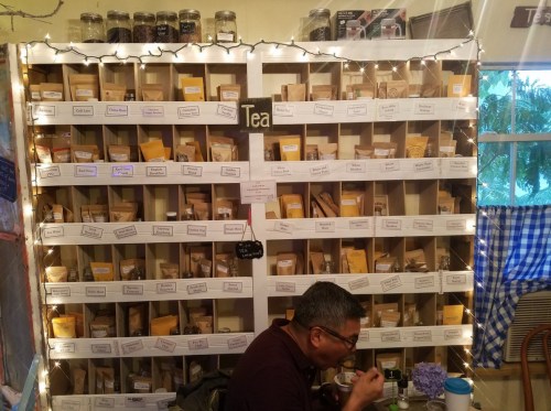 A cozy tea shop with a wall of neatly organized tea packets and a customer enjoying a meal at a table.