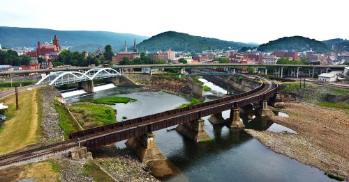 aerial of railroad tracks, a highway, river, and bridge with cumberland maryland and mountains in the background