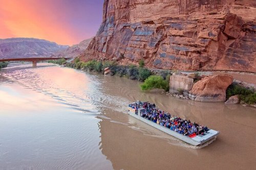 A large boat filled with people navigates a river surrounded by towering red rock cliffs at sunset.