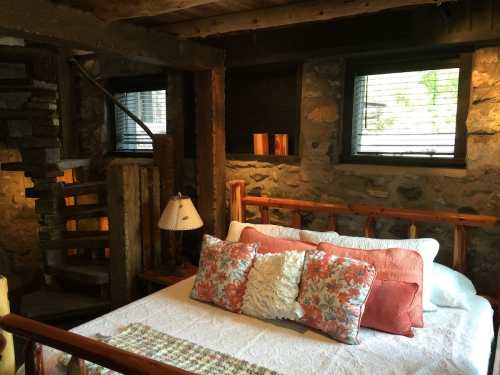Cozy bedroom with a stone wall, wooden bed, decorative pillows, and a spiral staircase in a rustic setting.