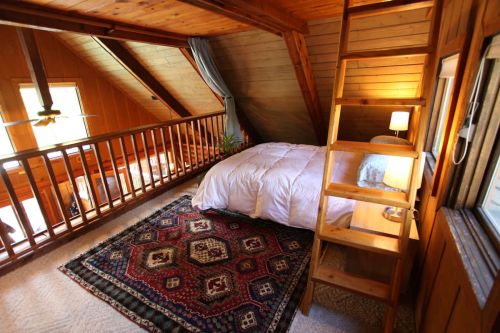 Cozy attic bedroom with wooden beams, a bed, a rug, and a small ladder leading to a loft area.