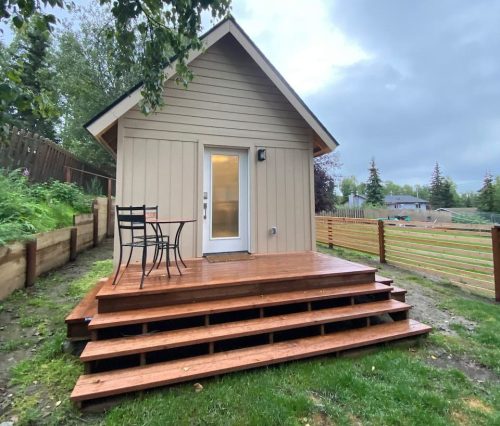 A small, light-colored cabin with a wooden deck and chairs, surrounded by grass and trees under a cloudy sky.