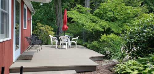 A cozy outdoor patio with a table and chairs, surrounded by greenery and a red umbrella, next to a red house.