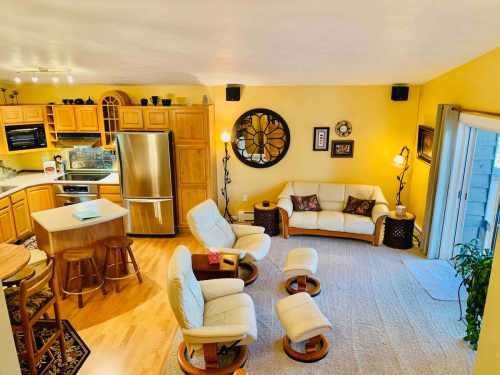 Cozy living room with yellow walls, white furniture, and a kitchen area, featuring a round clock and sliding glass door.