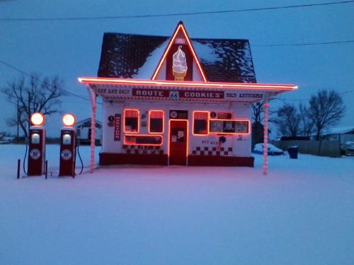 A retro ice cream shop on Route 66, illuminated with neon lights, surrounded by snow at dusk.