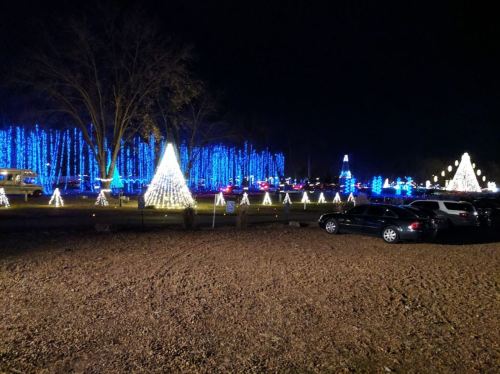 A festive display of blue and white Christmas lights, featuring illuminated trees and a parking area at night.