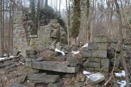 Ruins of a stone structure surrounded by trees and scattered snow, with crumbling walls and a rocky pathway.