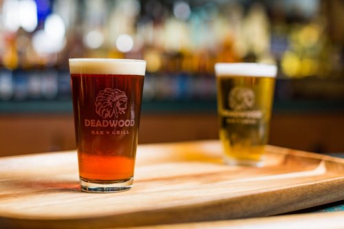 Two glasses of beer on a wooden tray, one dark and one light, with the logo of Deadwood Bar & Grill visible.