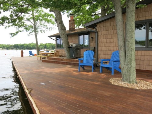 A wooden deck by a lake features blue chairs, a grill, and trees providing shade.