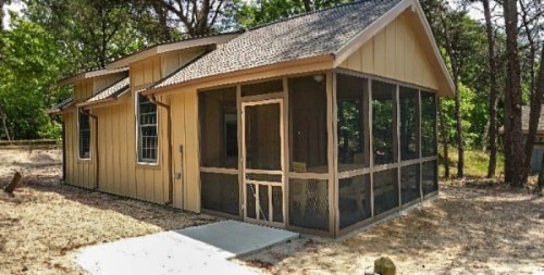 A small, yellow house with a screened porch, surrounded by trees and a sandy area.