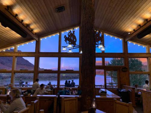 Cozy restaurant interior with wooden decor, large windows showcasing a scenic river view at sunset.