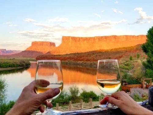 Two glasses of wine are raised in front of a scenic river and red rock formations at sunset.