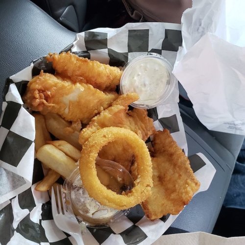 A takeout container with fried fish, onion rings, fries, and dipping sauce on a checkered paper.
