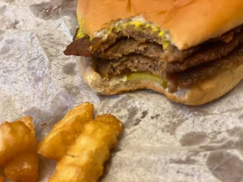 A partially eaten burger with multiple meat patties and mustard, alongside a few crinkle-cut fries on a paper wrapper.