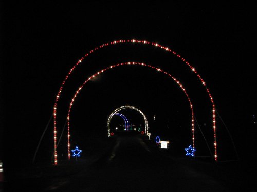 A dark road illuminated by colorful holiday lights forming arches and stars along the path.