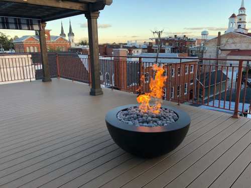 A rooftop terrace with a modern fire pit surrounded by stones, overlooking a cityscape at sunset.