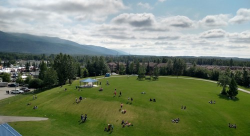 Aerial view of a green park with people relaxing, surrounded by trees and mountains under a partly cloudy sky.
