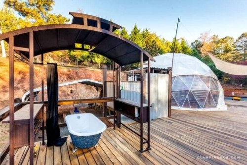 A wooden deck features a vintage bathtub and a geodesic dome structure in a forested outdoor setting.