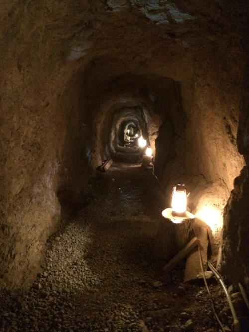 A dimly lit tunnel with stone walls, featuring lanterns on wooden stumps along a gravel path.