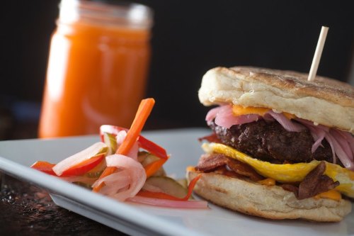 A close-up of a burger with bacon, cheese, and pickled onions, served with a side of colorful pickled vegetables and a drink.