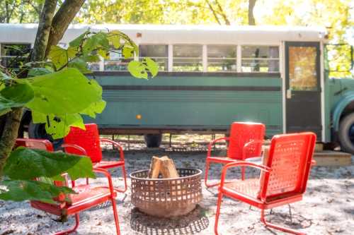 A vintage bus parked in a wooded area, surrounded by red chairs and a fire pit. Sunlight filters through the trees.