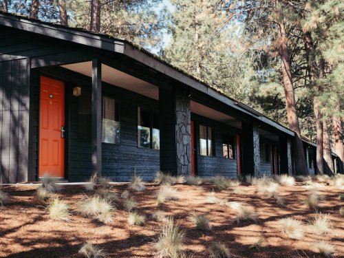 A row of black cabins with orange doors, surrounded by pine trees and grassy patches on a sunny day.