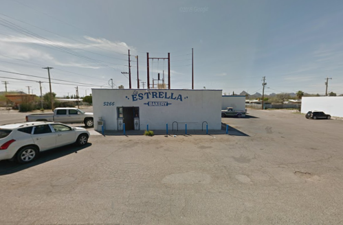 A white bakery building with "Estrella Bakery" painted on it, surrounded by parking and power lines under a clear sky.