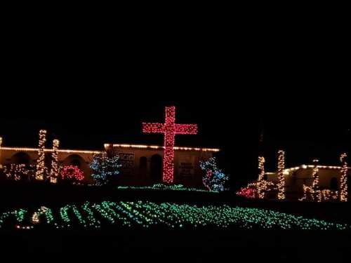 A brightly lit cross surrounded by colorful Christmas lights in a dark night setting.