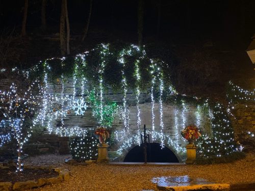 A stone archway adorned with cascading white lights and festive decorations, set in a dark, wooded area.