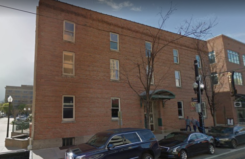A brick building with large windows and a green awning, parked cars in front, and trees nearby.