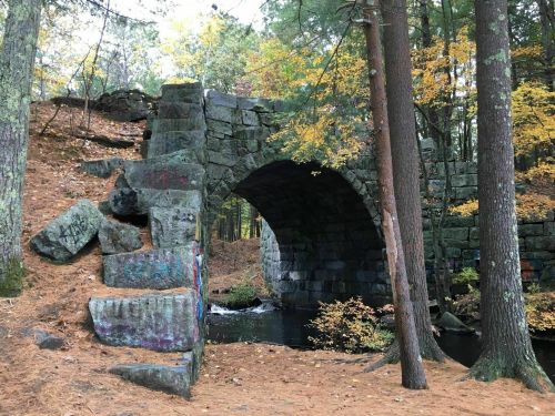 An old stone bridge arching over a small stream, surrounded by trees and autumn foliage.