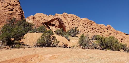 A rocky landscape featuring a natural arch, surrounded by shrubs and clear blue sky.