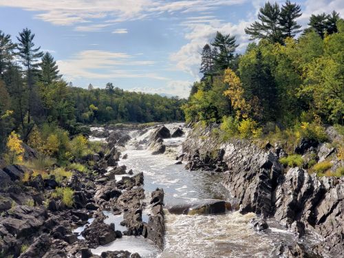 A scenic river flows through rocky terrain, surrounded by lush green trees and a clear blue sky with scattered clouds.