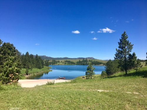A serene lake surrounded by green trees and hills under a clear blue sky with a few clouds.