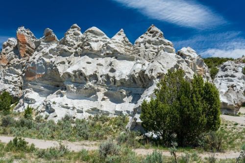 White rock formations with jagged peaks, surrounded by green shrubs and a blue sky with wispy clouds.