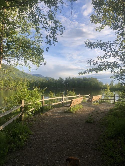 A serene lakeside view with benches, surrounded by lush greenery and mountains under a partly cloudy sky.