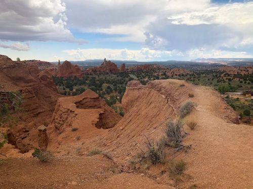 A rugged landscape with red rock formations and a winding dirt path under a cloudy sky.