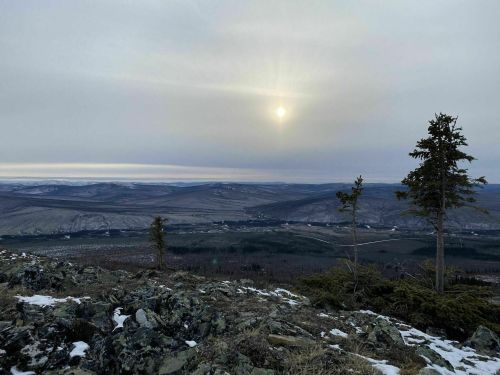 A panoramic view of a mountainous landscape under a cloudy sky, with the sun peeking through and sparse trees in the foreground.