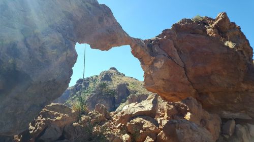 A natural rock arch stands against a clear blue sky, surrounded by rugged terrain and rocky formations.