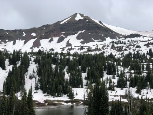 A snow-capped mountain rises above a forest of evergreen trees, reflecting in a serene lake under a cloudy sky.