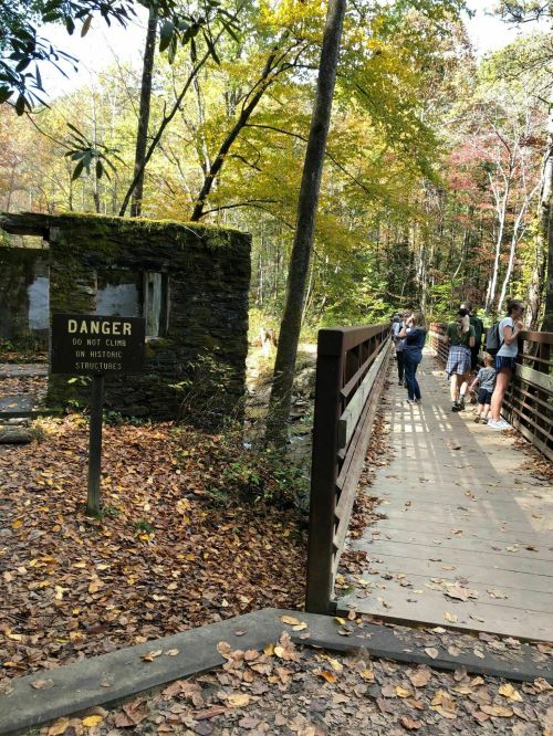 A wooden bridge leads to a stone structure with a warning sign about danger, surrounded by autumn foliage and visitors.