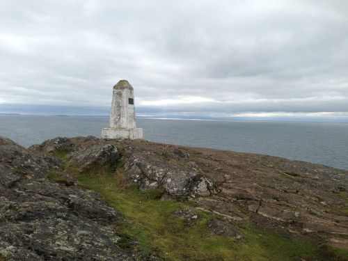 A white stone monument stands on rocky terrain overlooking a calm sea under a cloudy sky.