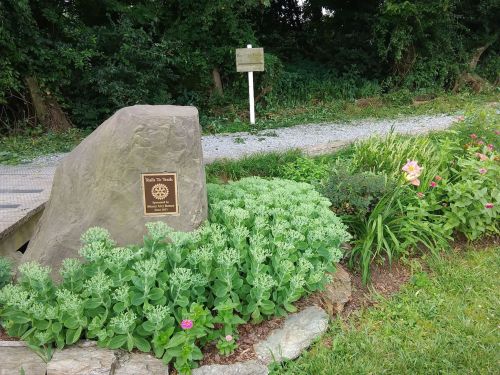 A stone monument with a plaque surrounded by green plants and flowers, set in a natural landscape.