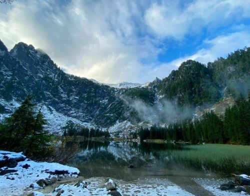 A serene mountain landscape with snow-capped peaks, a calm lake, and lush green trees under a cloudy sky.