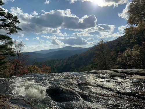 A scenic view of a flowing river with mountains and clouds in the background under a bright sky.